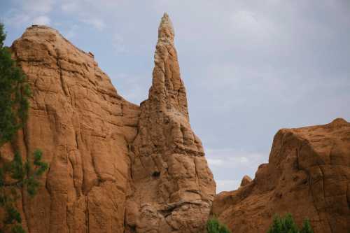 A tall, narrow rock formation rises from a rugged, reddish landscape under a cloudy sky.