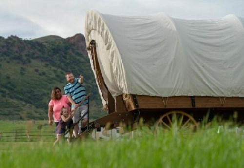 A family stands beside a covered wagon in a grassy field with rolling hills in the background.