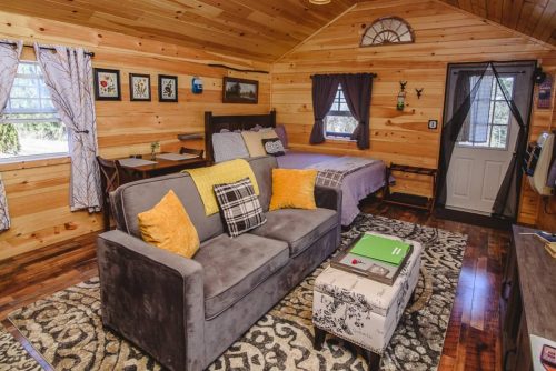 Cozy cabin interior with wooden walls, a bed, sofa with yellow pillows, and a door leading outside.