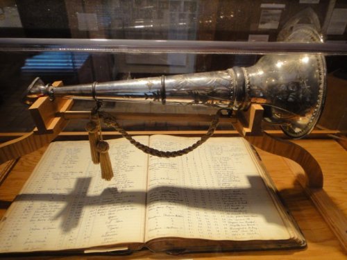 A silver trumpet displayed in a glass case, resting on an open book with handwritten entries.