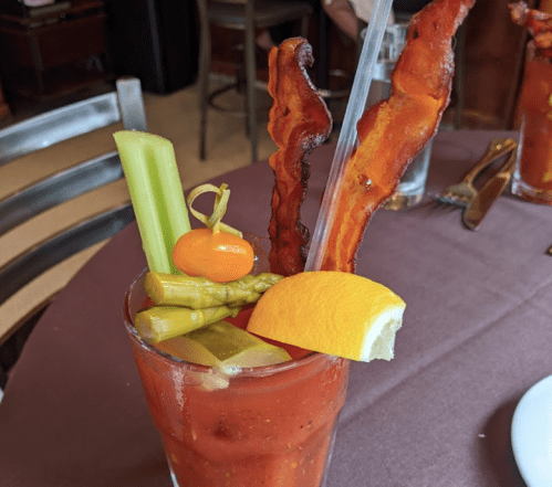 A vibrant Bloody Mary garnished with celery, asparagus, lemon, cherry tomato, and crispy bacon strips.