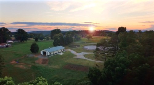 A scenic landscape at sunset, featuring a farm, fields, and distant hills under a colorful sky.