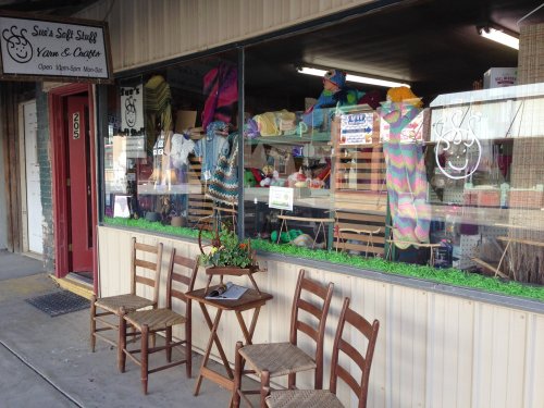 A craft store window displays colorful yarn and handmade items, with chairs outside for visitors to sit.