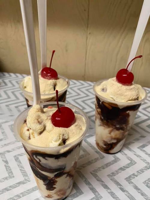 Three cups of ice cream topped with whipped cream and cherries, served with plastic spoons on a patterned table.