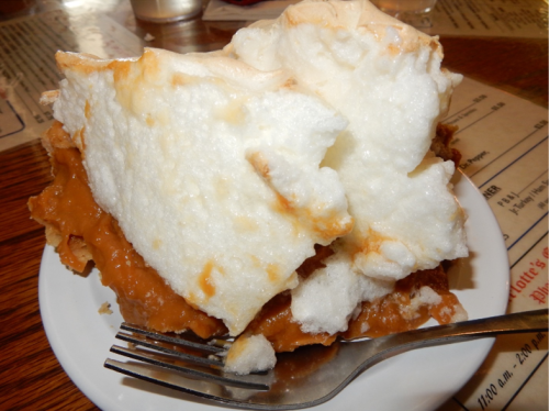 A slice of pie with a fluffy meringue topping and a creamy filling on a white plate, with a fork beside it.