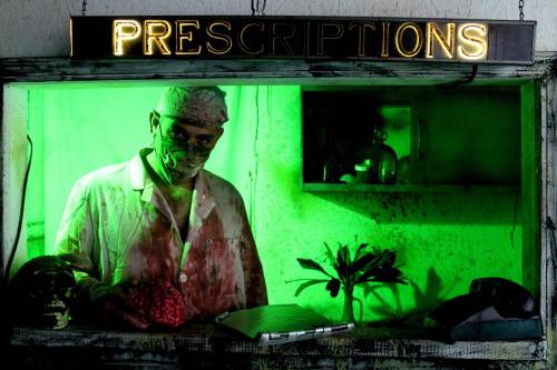 A creepy figure in a blood-stained outfit stands behind a glowing "Prescriptions" sign in a dimly lit setting.