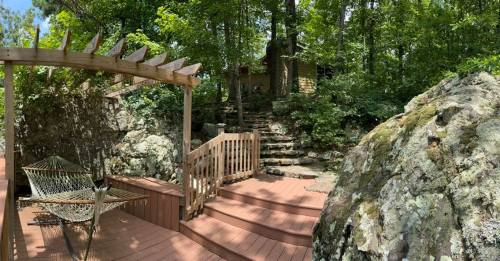 A wooden deck with a hammock, surrounded by lush greenery and rocks, leading to a stone staircase in a serene outdoor setting.