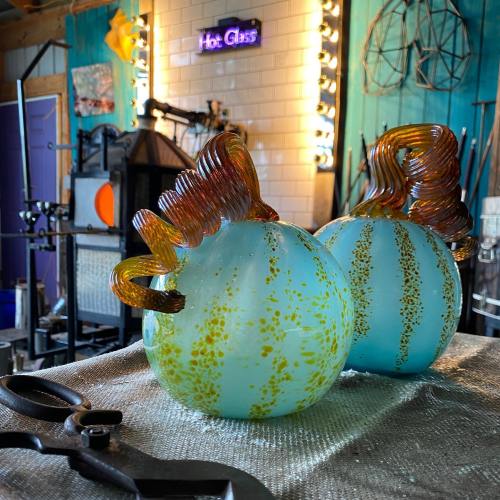 Two colorful glass art pieces with swirled patterns, set in a bright workshop with tools and lights in the background.