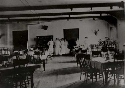 A vintage dining room with wooden tables and chairs, featuring staff in white uniforms and a rustic decor.