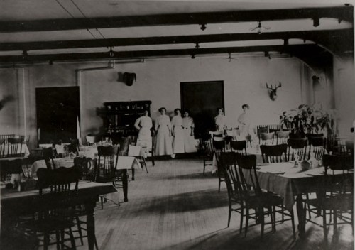 A vintage dining room with wooden tables and chairs, featuring staff in white uniforms and a rustic decor.
