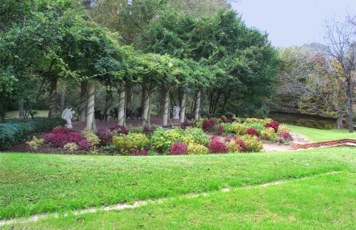 A serene garden with lush greenery, colorful flowers, and a colonnade featuring sculptures in the background.