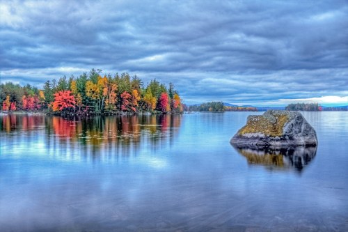 A tranquil lake scene with colorful autumn trees reflecting on the water under a cloudy sky. A large rock is in the foreground.