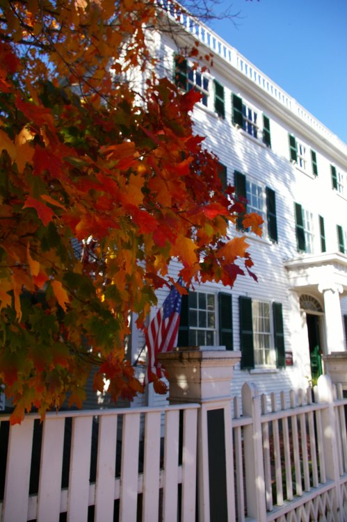 A white building with green shutters, framed by vibrant autumn leaves and an American flag, under a clear blue sky.