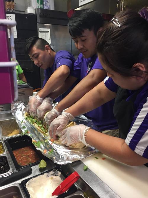 Three people in purple shirts prepare large sandwiches in a busy kitchen, wearing gloves and working together.