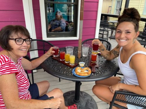 Two women sit at a table with colorful drinks and a pastry, smiling and enjoying their time together outdoors.
