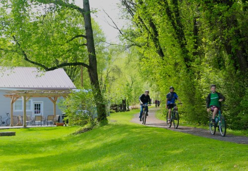 Three people ride bicycles along a path beside a house surrounded by lush greenery on a sunny day.