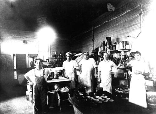A group of five men in aprons stand in a vintage kitchen, surrounded by cooking equipment and ingredients.