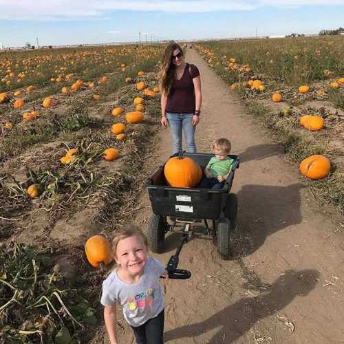 A woman stands in a pumpkin patch while a girl pulls a wagon with a child and a large pumpkin.