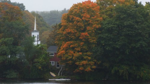 A serene lakeside scene with colorful autumn trees, a church steeple, and a house partially hidden by foliage.