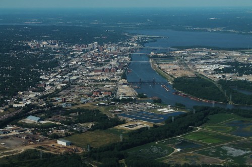 Aerial view of a city with a river, industrial areas, and green spaces under a clear blue sky.