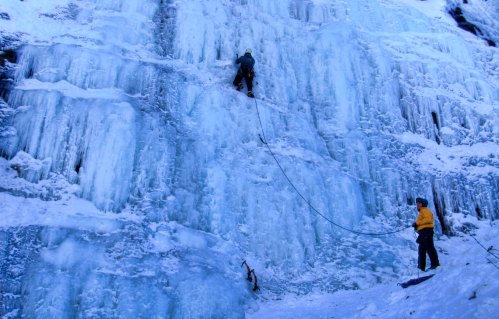Two climbers ascend a frozen ice wall, one climbing while the other secures the rope below.