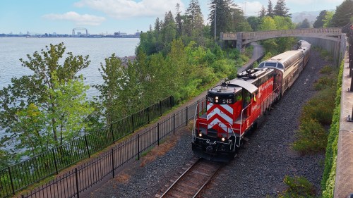 A red and white train travels along a scenic waterfront track, surrounded by trees and a calm body of water.