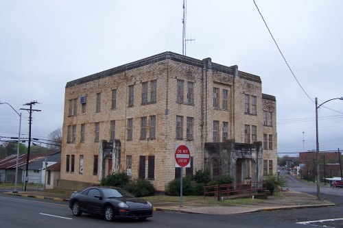 Abandoned two-story building with a weathered facade, surrounded by overgrown plants and a nearby stop sign.
