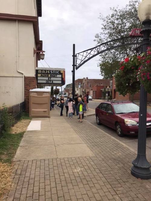A group of people stands outside the Majestic Movie Theater on a cloudy day, with a red car parked nearby.
