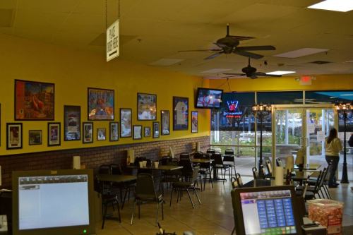 Interior of a casual dining restaurant with yellow walls, tables, and a TV, featuring framed artwork and a view of the entrance.