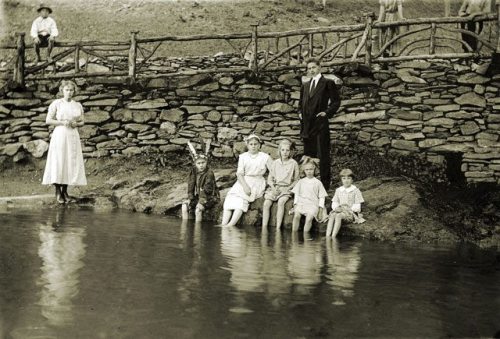A vintage photo of a group of children and adults by a pond, with a stone wall in the background.