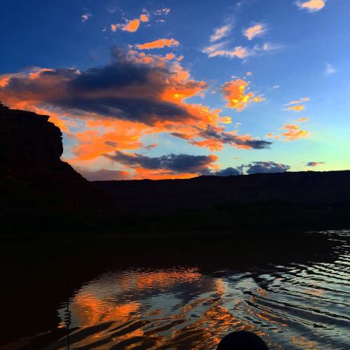 A vibrant sunset reflects on calm water, with dramatic clouds and rocky terrain in the background.