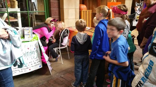 A group of children wait in line for face painting at a festive event, with colorful tables and decorations in the background.