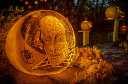 A carved pumpkin featuring a portrait of Shakespeare, surrounded by autumn leaves and glowing jack-o'-lanterns.