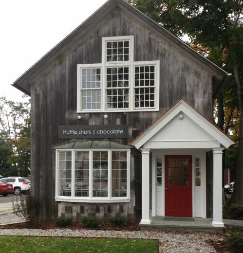 A charming wooden building with a red door, featuring a sign that reads "truffle shots | chocolate." Green lawn and gravel path.