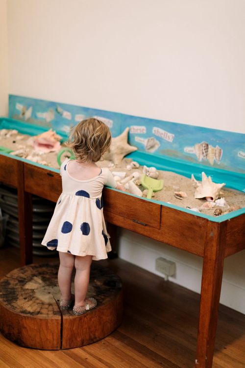 A child in a polka dot dress explores a sensory table filled with sand and seashells.