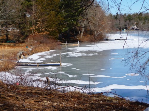 A frozen lake with patches of ice and a wooden dock, surrounded by bare trees and winter scenery.
