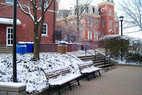 Snow-covered benches in a park-like area, with a historic building and trees in the background.