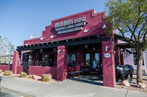 Exterior of a red adobe-style restaurant with a sign, surrounded by desert landscaping and a decorative horse statue.