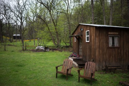 A small wooden cabin surrounded by trees, with two wooden chairs in front and a grassy area.