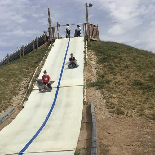 Two children slide down a large, sloped slide on a grassy hill, with a few people standing at the top.