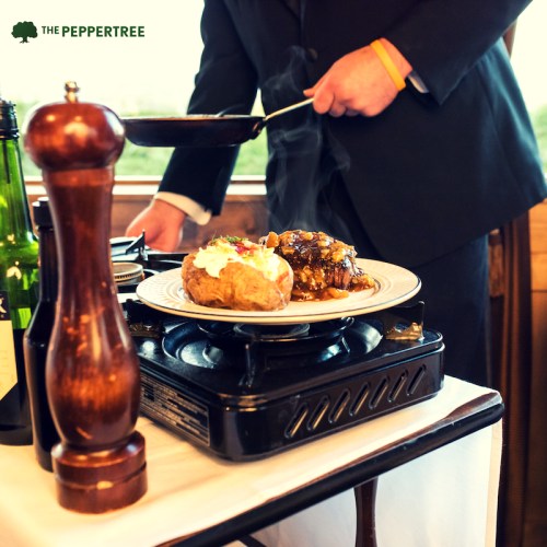 A chef in a suit prepares a plate of food on a portable stove, with a pepper grinder and drinks nearby.