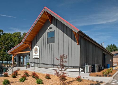 A modern brewery building with a wooden roof, large windows, and landscaped surroundings under a clear blue sky.
