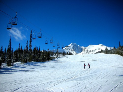Two skiers on a snowy slope with ski lifts and pine trees under a clear blue sky.
