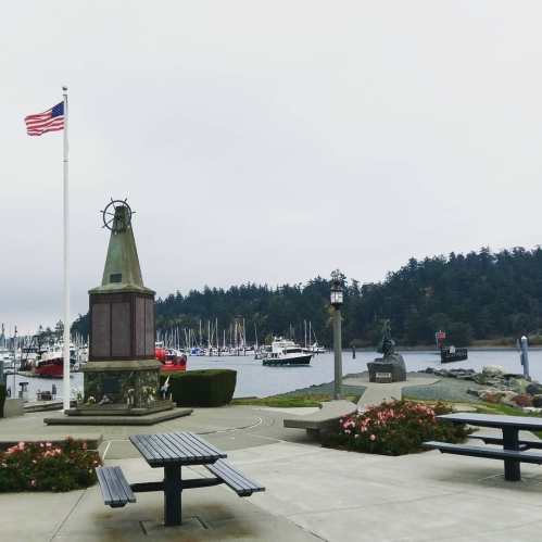 A waterfront park with a flag, monument, benches, and boats in the harbor under a cloudy sky.