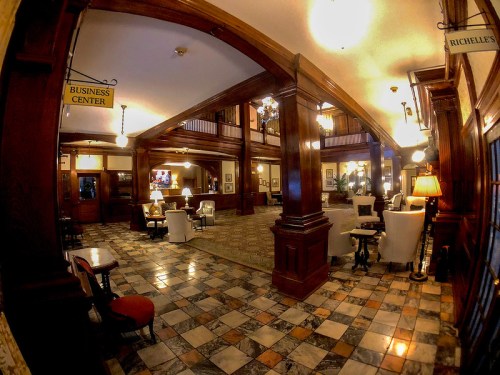 A spacious hotel lobby with wooden pillars, elegant seating, and a business center sign, featuring warm lighting and patterned flooring.