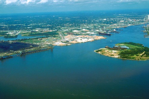 Aerial view of a river delta with industrial areas, ships, and green land along the water's edge under a cloudy sky.