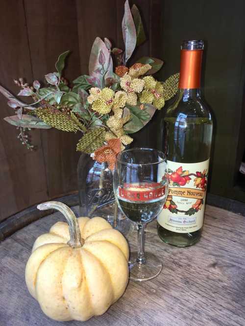 A bottle of Pomme Nouveau wine, a glass, a yellow pumpkin, and a floral arrangement on a wooden surface.