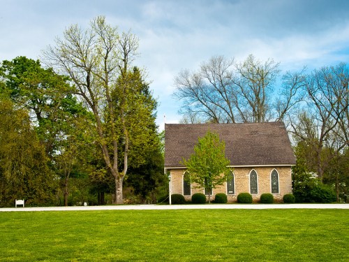 A quaint stone church with arched windows, surrounded by lush greenery and trees under a cloudy sky.