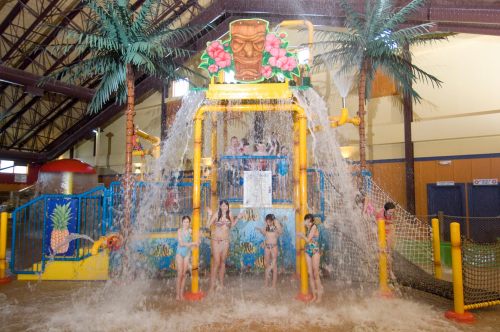 Children play under a large water feature with palm trees in an indoor water park, enjoying splashes and fun.
