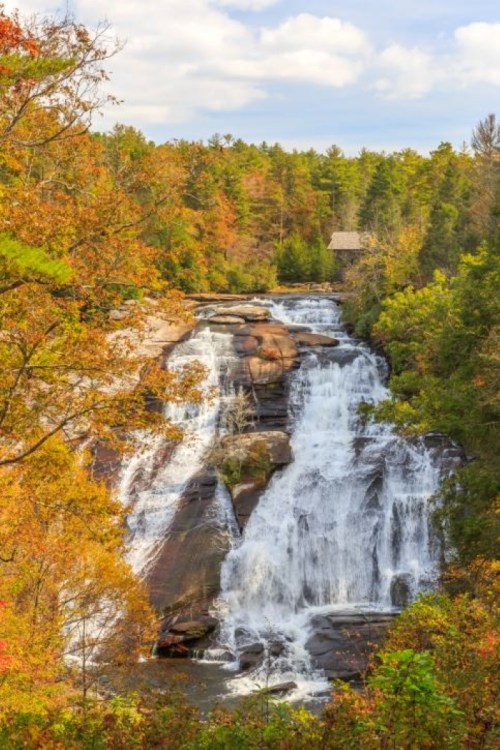 A scenic waterfall surrounded by vibrant autumn foliage and a distant cabin under a partly cloudy sky.